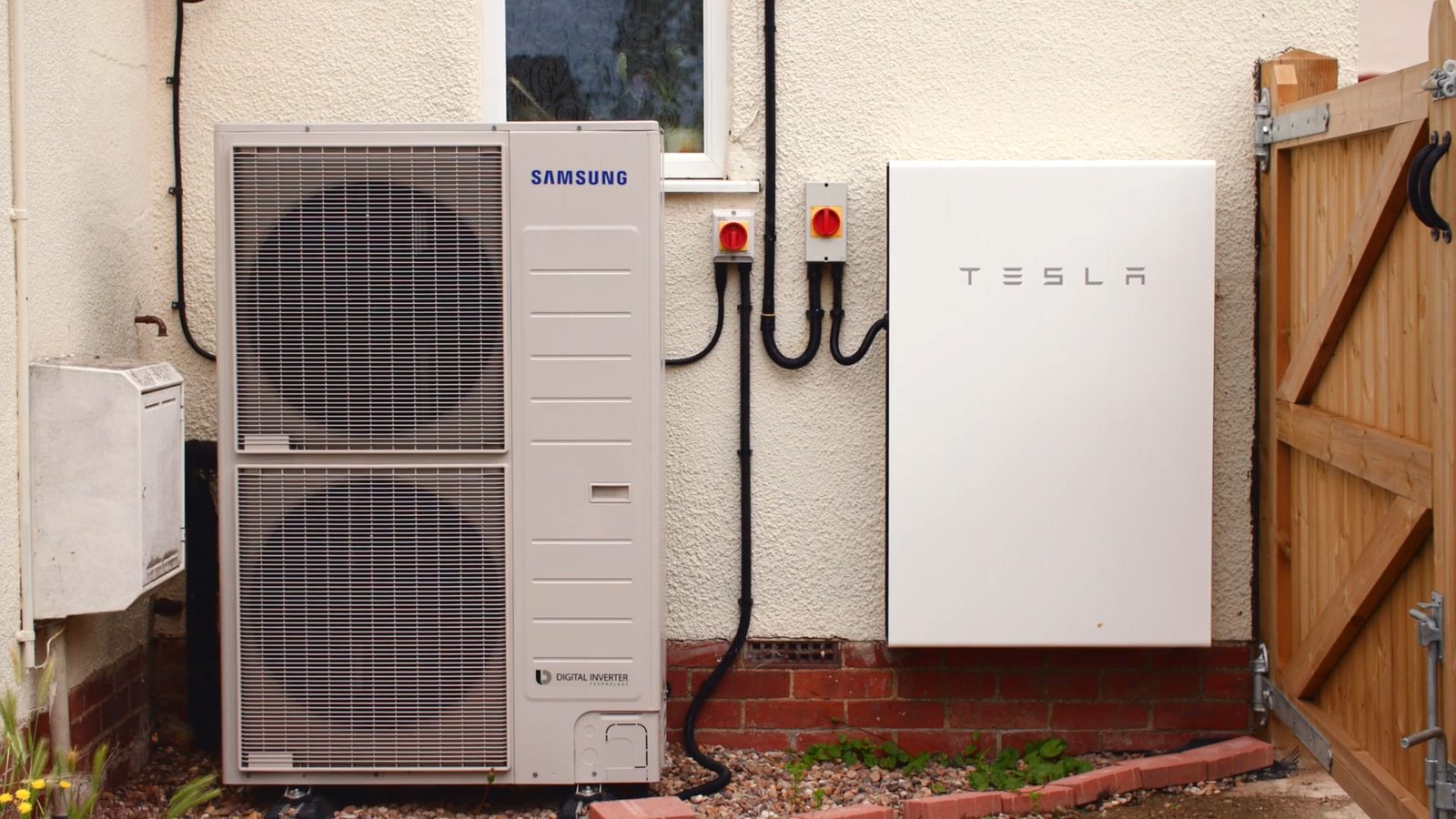 Buitenunit van een warmtepomp en een witte thuisbatterij naast elkaar tegen de gevel van een woning, naast een houten poort