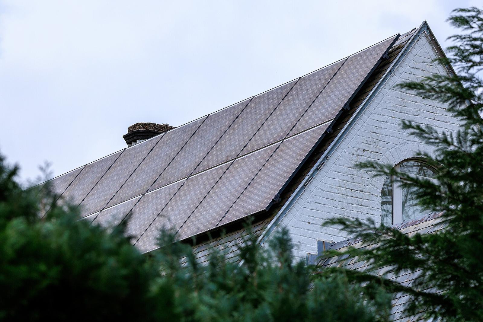 Schuin dak volledig bedekt met zwarte zonnepanelen boven een witgeschilderde bakstenen puntgevel, achter groene coniferen